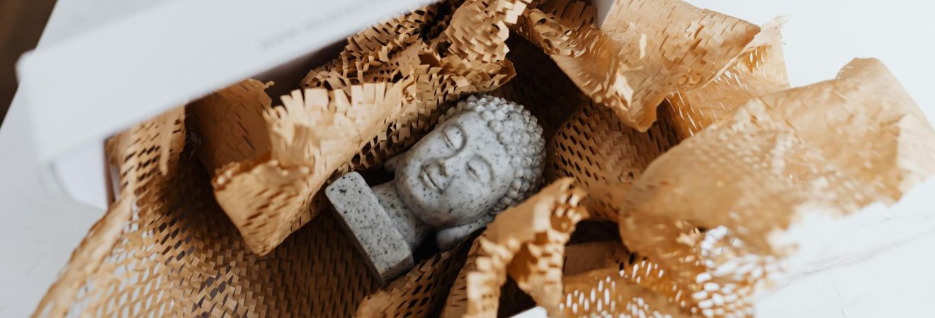 A small stone Buddha statue nestled in crumpled kraft paper inside an open white gift box on a tabletop.