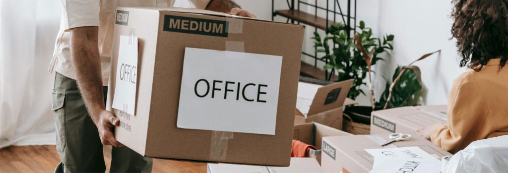 Person carrying a labeled “OFFICE” box during IT office relocation in a bright workspace with packing materials